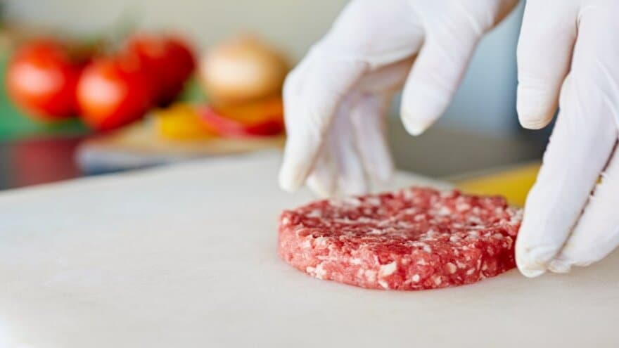 Hender med hvite plasthansker behanlder rå hamburger