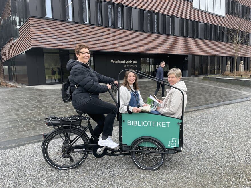 Tre bibliotekarer leser bøker om bærekraft mens de sykler rundt med el-sykkel på campus NMBU
